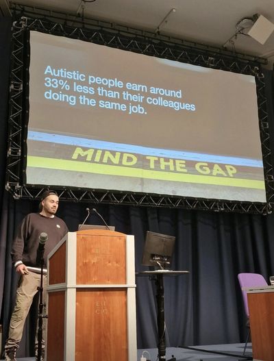 Craig speaks beside the lectern at Accessibility Scotland, under a slide stating "Autistic people earn around 33% less than their colleagues doing the same job" above a photo of a "MIND THE GAP" platform edge.