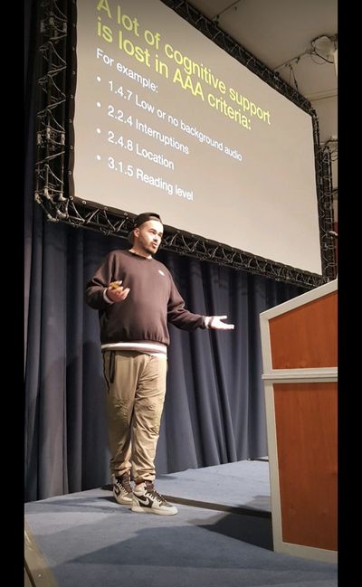 Craig spreads his arms mid-explanation at Accessibility Scotland, in front of a slide headed "A lot of cognitive support is lost in AAA criteria".