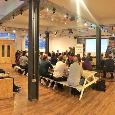 A packed room at Campus North in Newcastle, with the audience seated at white picnic-style benches while Craig presents in the far corner.