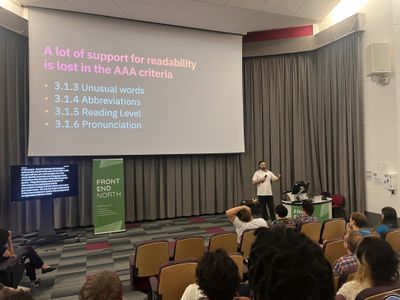 Craig gestures towards a slide titled "A lot of support for readability is lost in the AAA criteria", listing unusual words, abbreviations, reading level and pronunciation, with live captions on a screen beside the stage.