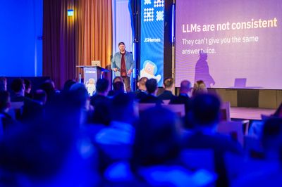 Craig walks the JSHeroes stage in front of a slide stating "LLMs are not consistent — they can't give you the same answer twice."