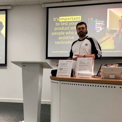 Craig stands behind a desk mid-talk, in front of a slide reading "It's important to test your product with people who use assistive technology".