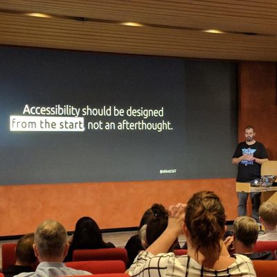 Craig on stage in front of a seated audience, presenting a slide reading "Accessibility should be designed from the start, not an afterthought".