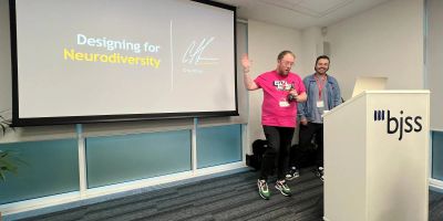 Craig waves to the audience at A11y North in a pink a11y North t-shirt, as his "Designing for Neurodiversity" title slide fills the screen.