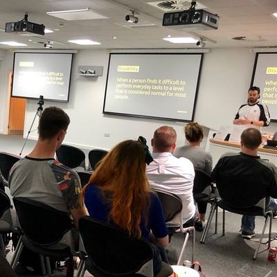 Craig presents to a seated audience across three screens, each showing a slide defining disability as "when a person finds it difficult to perform everyday tasks to a level that is considered normal for most people".