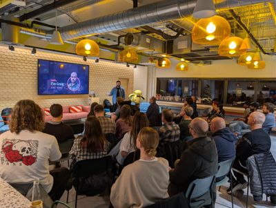 Craig introduces himself at a cosy NUX meetup under amber pendant lamps, with his "Hello, I'm Craig" slide showing ADHD and Autistic badges on a wall-mounted screen.