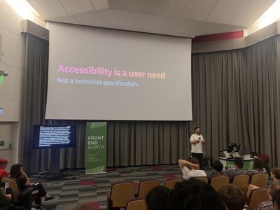 Craig smiles at the Front End North audience in front of a slide reading "Accessibility is a user need — not a technical specification", with live captions displayed on a side screen.