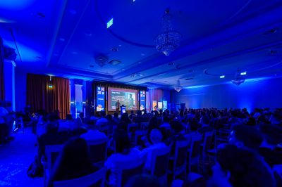 A wide view of the chandelier-hung JSHeroes ballroom bathed in blue light, with Craig on the distant stage presenting Deque's claim that accessibility work can be done with zero specialised accessibility knowledge.