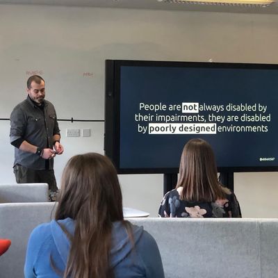 Craig speaks to a small workshop audience beside a screen reading "People are not always disabled by their impairments, they are disabled by poorly designed environments".