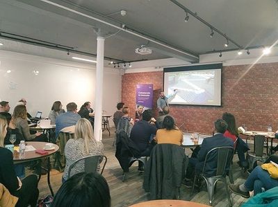 A wide view of a full meetup room, with Craig presenting beside a DWP Digital "Collaborate to innovate" banner at the front.