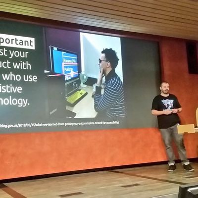 Craig on stage beside a slide reading "It's important to test your product with people who use assistive technology", showing a photo of a screen reader user at their desk.