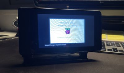 A Raspberry Pi with a 7 inch touchscreen on a desk stand, booting Raspberry Pi OS and showing the Welcome to the Raspberry Pi Desktop splash screen.