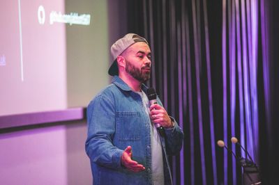 Craig gestures while speaking into a microphone at Access Given, wearing a backwards cap and denim shirt.