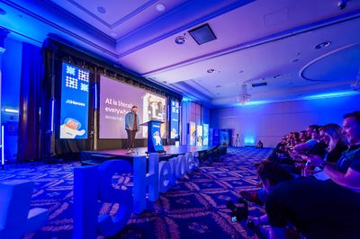Craig smiles on the JSHeroes stage in a blue-lit ballroom, beside a slide reading "AI is literally everywhere. Annoyingly…", with giant JSHeroes letters at the stage edge.