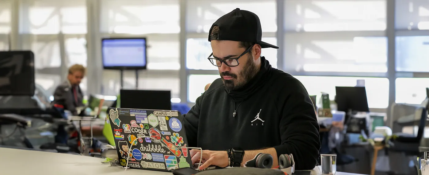 A white man with a short black beard, He is wearing glasses and a black backwards baseball cap. He is typing on a laptop covered with stickers in an open plan office.